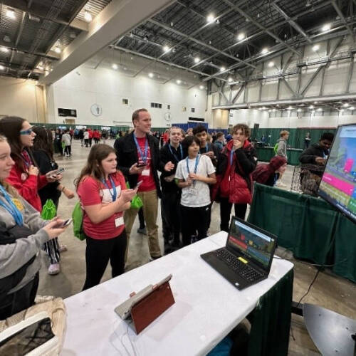 A group of people gathered around a table with a laptop and a large screen displaying a colorful quiz question in an indoor event space.